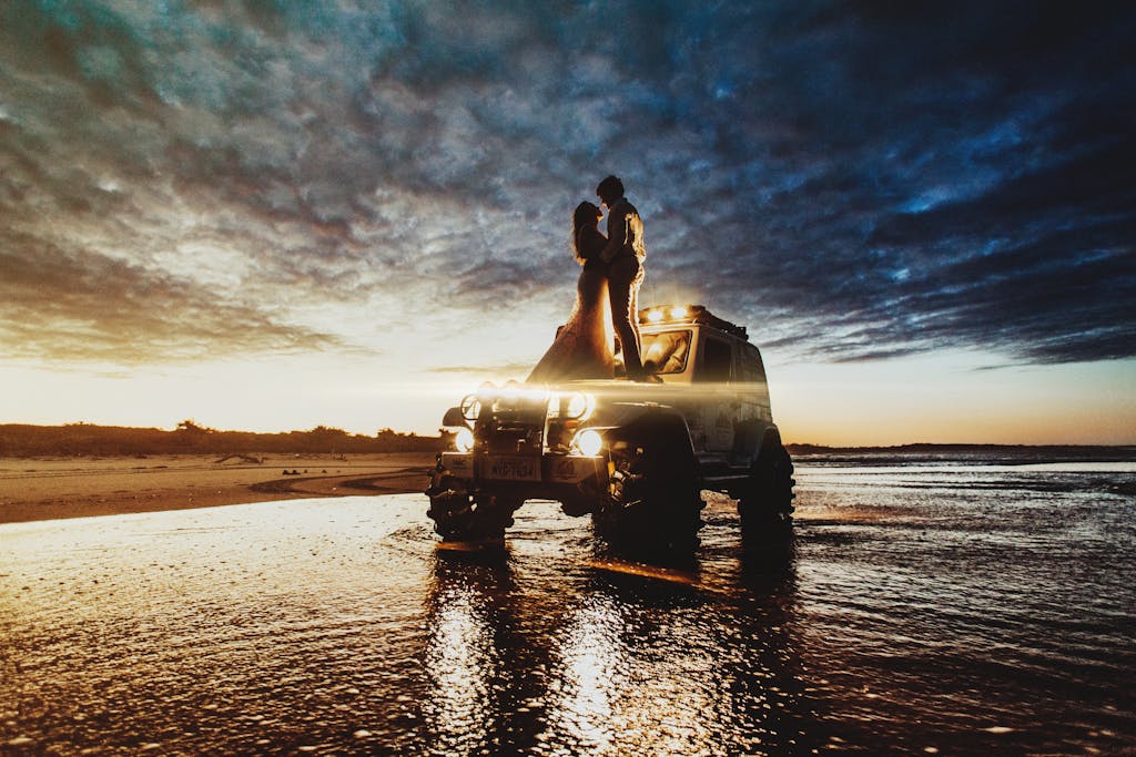 Couple embracing atop a 4x4 vehicle during a dramatic sunset on a beach.